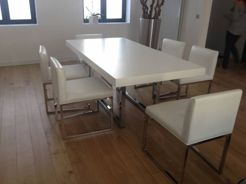 Salle à manger moderne avec grande table blanche et huit chaises rembourrées claires, sur parquet. Fenêtres lumineuses.