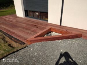 Terrasse en bois exotique rougeâtre avec jardinière triangulaire en gravier, bordant une façade blanche et un volet gris. REDMI NOTE 8 AI QUAD CAMERA.