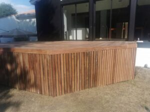 Terrasse moderne en bois exotique, avec jupe à lattes verticales, devant une maison contemporaine avec grandes baies vitrées.