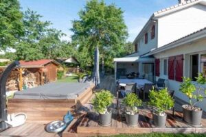 Spacieux jardin aménagé avec grande terrasse en bois, jacuzzi couvert, salon de jardin sous pergola. Maison blanche aux volets rouges.