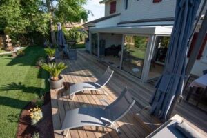 Terrasse en bois ensoleillée avec chaises longues grises et plantes, bordant une véranda vitrée et un jardin verdoyant.
