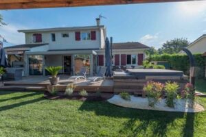 Grande terrasse en bois avec véranda, salon de jardin et jacuzzi intégré dans un jardin verdoyant. Maison moderne à volets rouges.