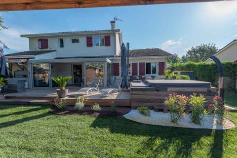 Grande terrasse en bois avec véranda, salon de jardin et jacuzzi intégré dans un jardin verdoyant. Maison moderne à volets rouges.