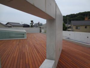 Grande terrasse de toit moderne en bois avec murs et poutre en béton. Puits de lumière vitré, vue sur des maisons et une colline verdoyante.