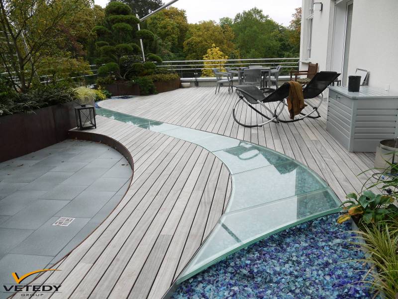 Terrasse de toit moderne en bois clair avec un chemin vitré incurvé sur des roches bleues. Espace salon, salle à manger et jardinières. VETEDY GROUP.