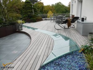 Terrasse de toit moderne en bois clair avec un chemin vitré incurvé menant à un lit de verre bleu. Aménagements paysagers luxuriants et mobilier extérieur.