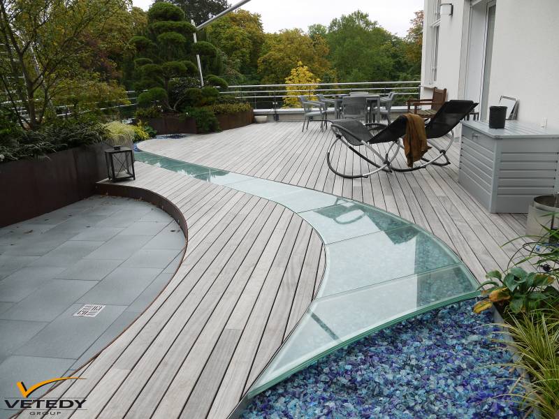 Terrasse de toit moderne en bois clair avec un chemin vitré incurvé menant à un lit de verre bleu. Aménagements paysagers luxuriants et mobilier extérieur.