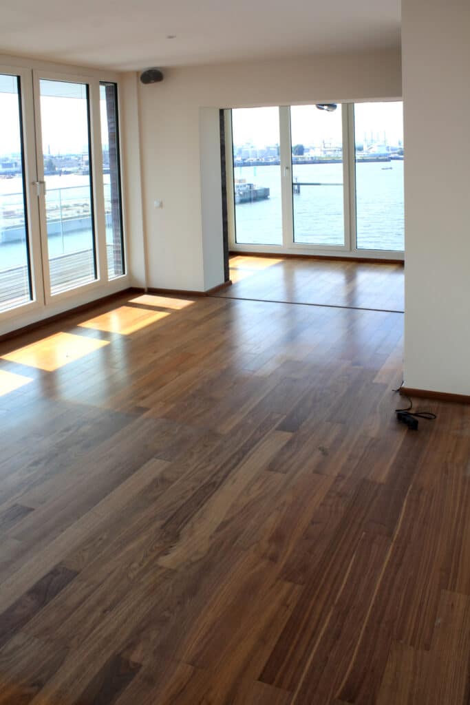 Grand salon vide avec parquet moderne en bois et grandes fenêtres offrant une vue imprenable sur un port animé, baigné de lumière naturelle.