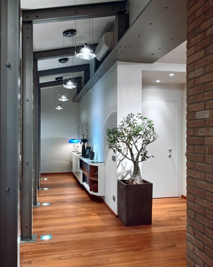 Couloir moderne avec plancher bois, poutres métalliques exposées, suspensions design, console blanche et plante. Lumières au sol bleues.