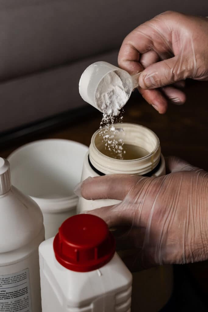 Main gantée versant de la poudre blanche d'une cuillère doseuse dans un récipient. Bouteilles chimiques, texte 'orina' visible.