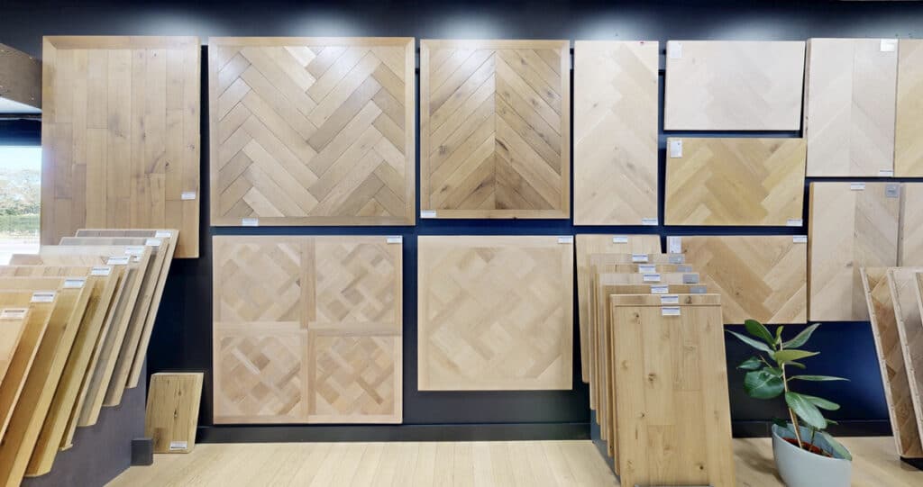 Parquet bois clair: échantillons, chevrons & motifs Showroom de parquets en bois clair: grands échantillons muraux (chevrons, lames droites, motifs variés) et dalles sur présentoirs. Plante verte.