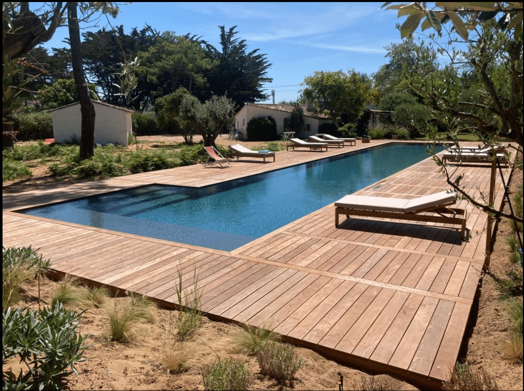 Piscine moderne avec terrasse bois : Luxe et détente. Grande piscine rectangulaire entourée d'une vaste terrasse en bois clair avec chaises longues, sous un ciel bleu estival, dans un jardin.