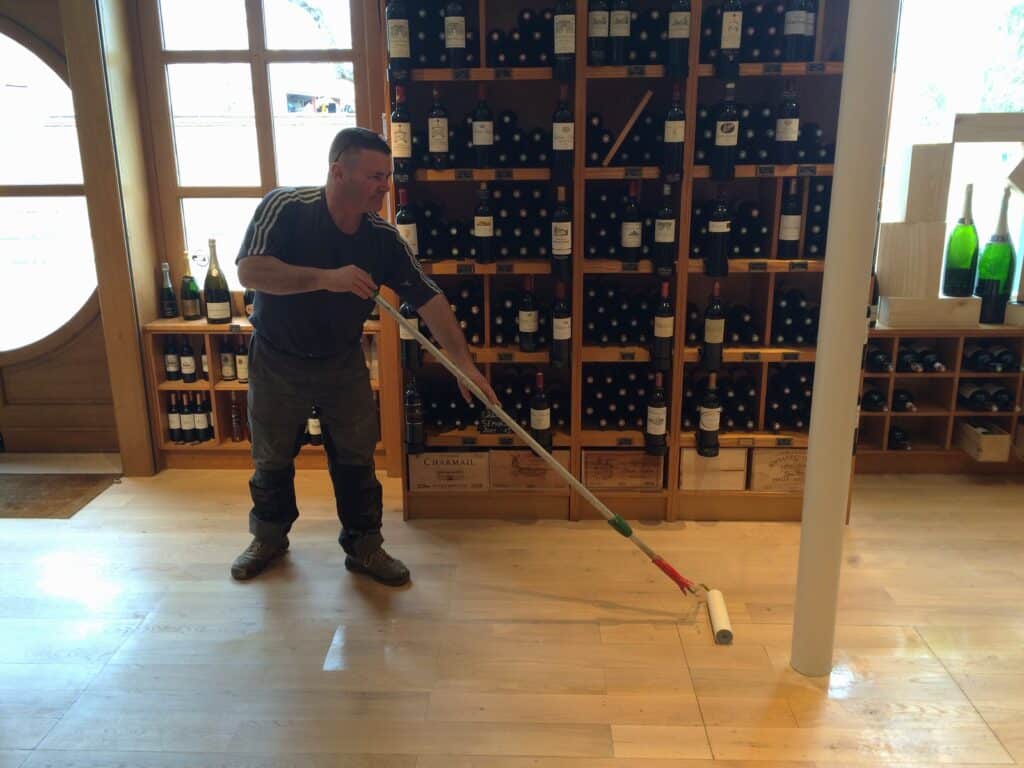 Rénovation parquet cave à vin : application vernis Homme appliquant du vernis sur un parquet en bois clair avec un rouleau. Au fond, un grand casier à vin rempli de bouteilles, dont des boîtes "CHARMAIL".