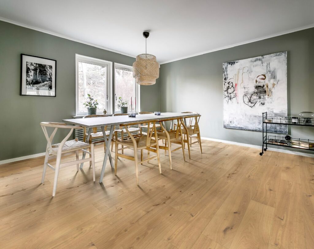 Salle à manger moderne : murs verts, table bois clair, parquet Salle à manger moderne aux murs verts, table blanche, chaises en bois clair, parquet clair. Suspension tressée, tableaux muraux.