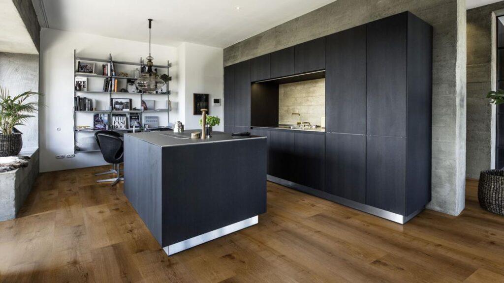 Cuisine ouverte noire minimaliste, îlot central foncé, et armoires intégrées. Planchers en bois chaud contrastant avec le béton industriel.
